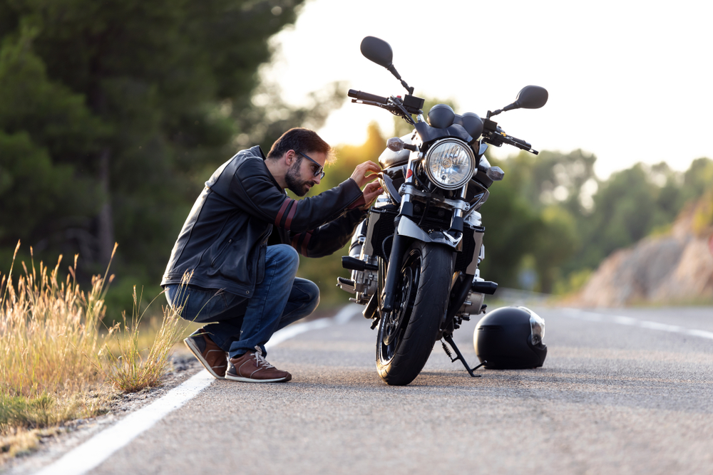 A man squatting down next to his motorcycle parked on the side of a road checking the engine.