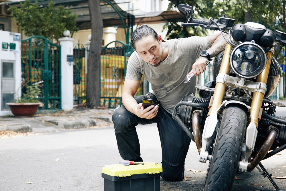 A man kneeling next to his motorcycle holding tools and looking at the engine.
